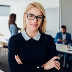 Smiling professional wearing glasses and blue sweater in meeting room with colleagues in the background