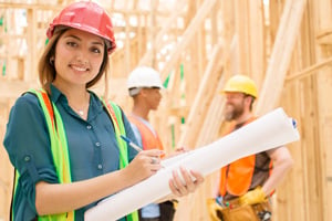 Construction manager at job site wearing hard hat and safety vest holding blueprints and smiling as colleagues talk in the background