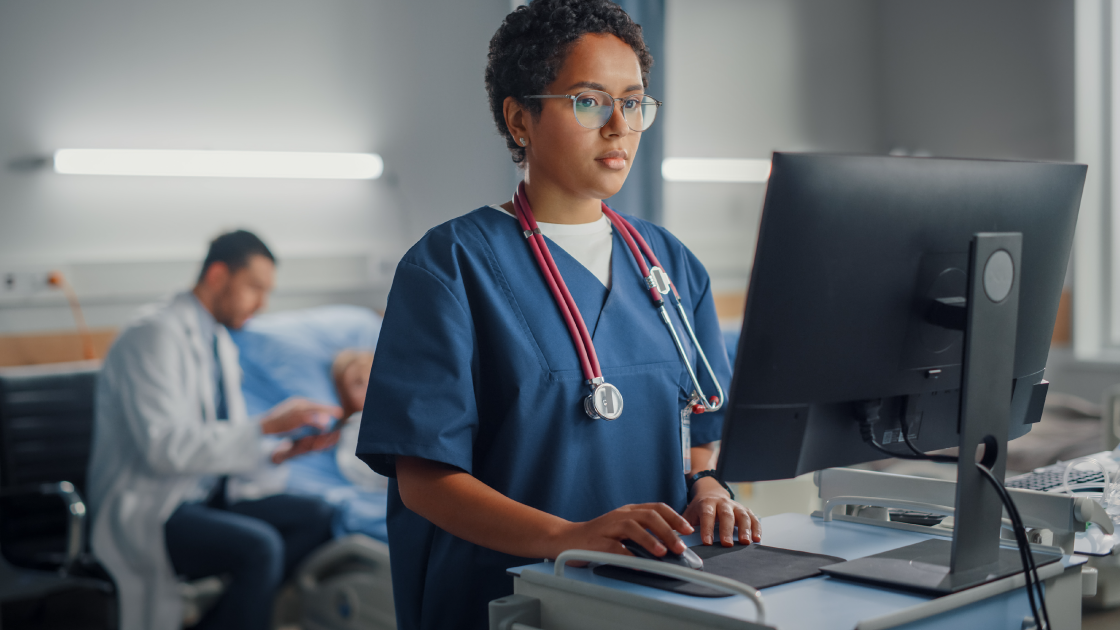 Woman in scrubs with a stethoscope around her neck is at a computer in a medical room.