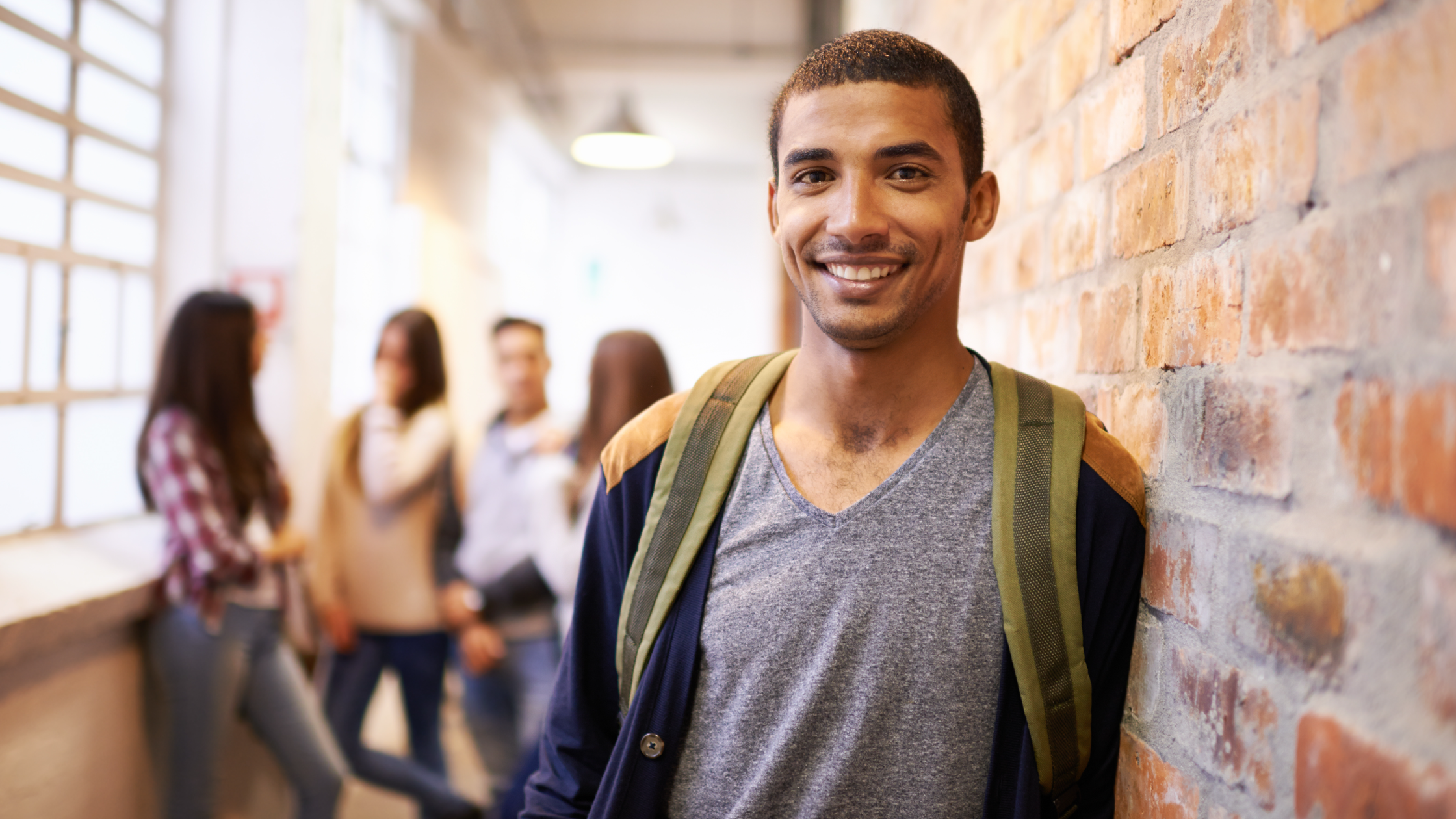 Smiling college student with backpack standing in brick hallway with classmates in background