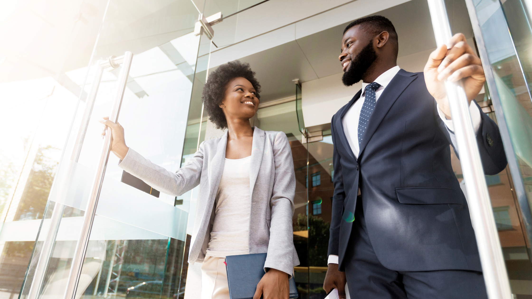 Professional business colleagues in formal attire exit modern glass office building in bright daylight