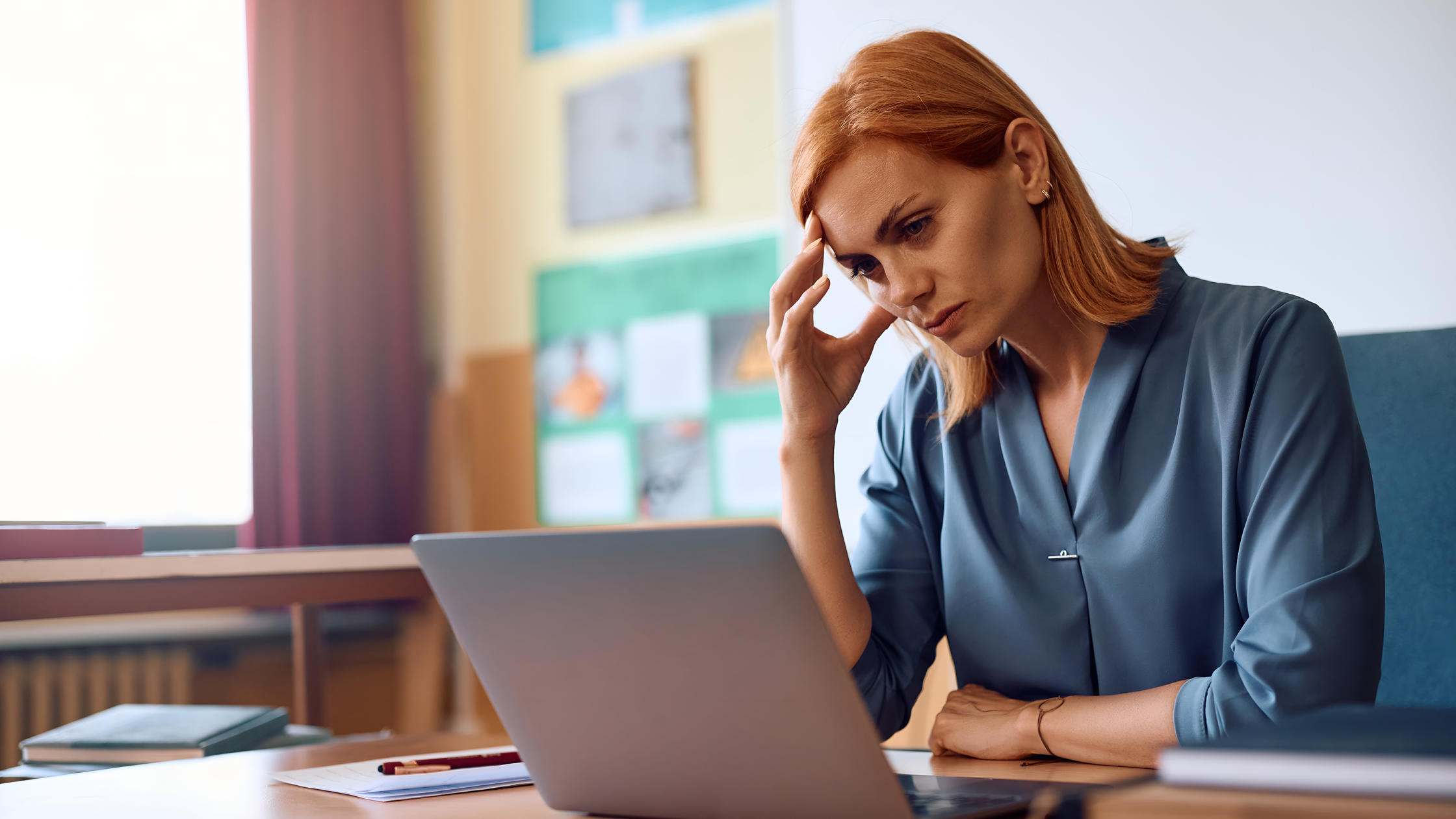 Professional woman with red hair concentrating on laptop work with hand on forehead in bright office