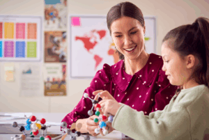 Smiling science teacher in polka dot blouse works with student using colorful molecular models in bright classroom