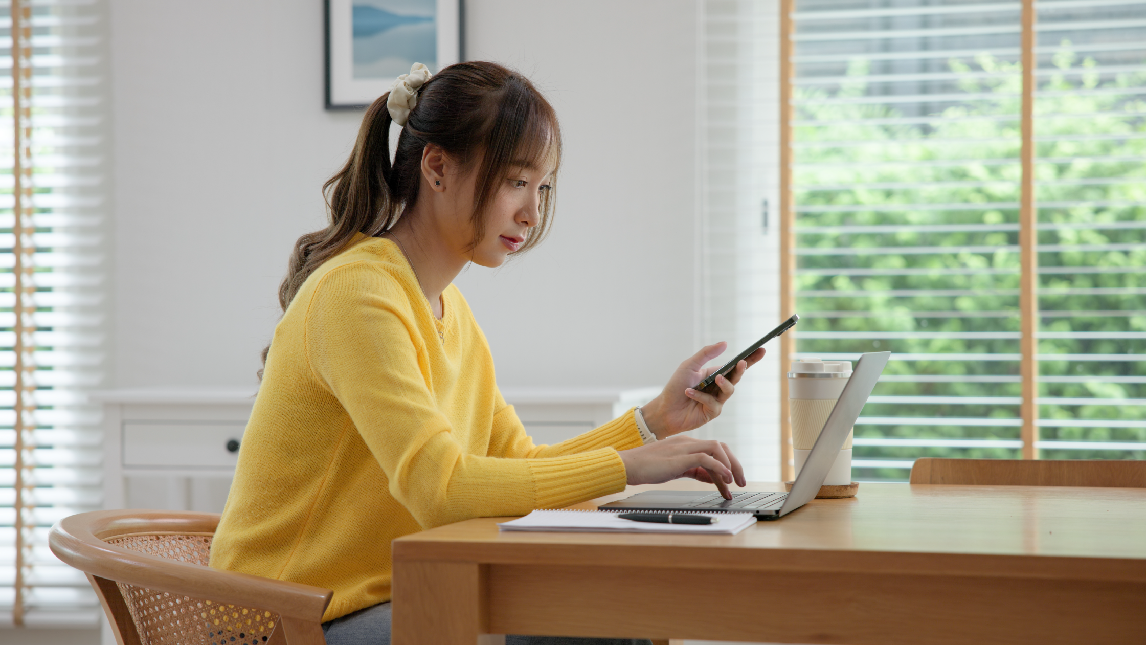 Woman in a yellow blouse looks at her phone and computer in the office.