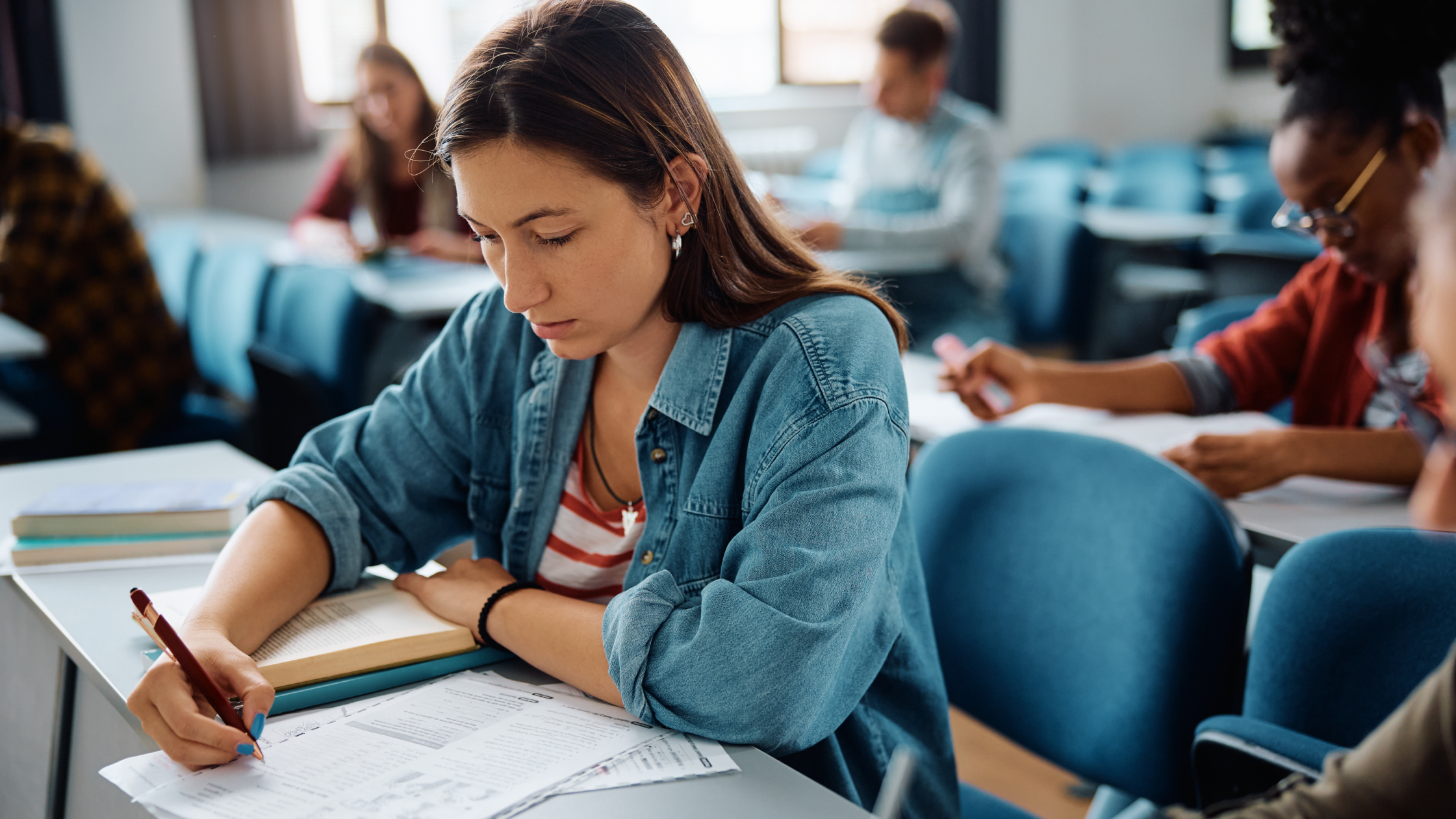 Female college student taking exam in classroom with classmates studying in background