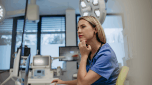 Female nurse in blue scrubs looking thoughtfully at computer screen in modern hospital examination room