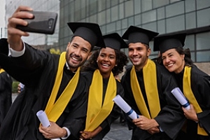 Four MBA students wearing black caps and gowns with gold neck sashes take a selfie together at graduation